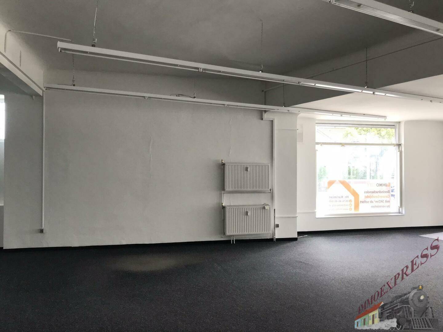 Innenansicht des Geschäftsraums mit Teppichboden, Heizkörpern und einem großen Fenster.