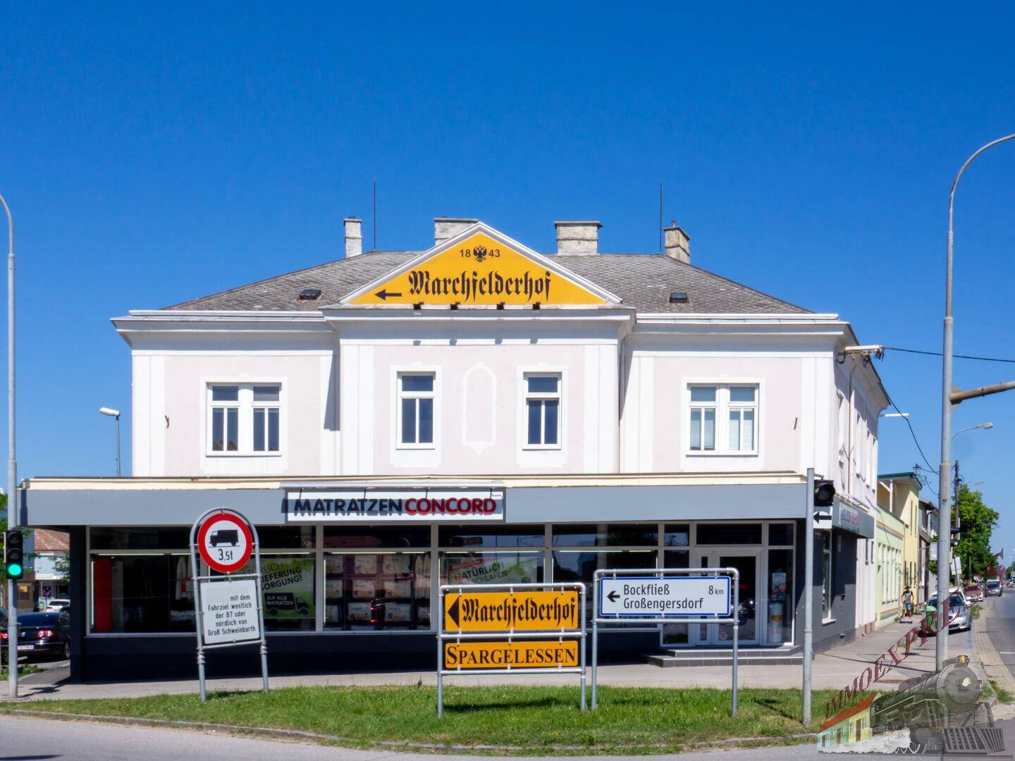 Ansicht des historischen Gebäudes "Marchfelderhof" mit Geschäften im Erdgeschoss und Straßenschildern.