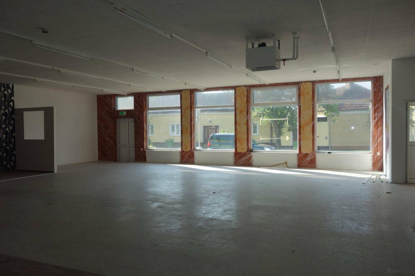 Geräumiger Gewerberaum mit großen Fenstern zur Straße und Fliesenboden, der viel Tageslicht erhält.