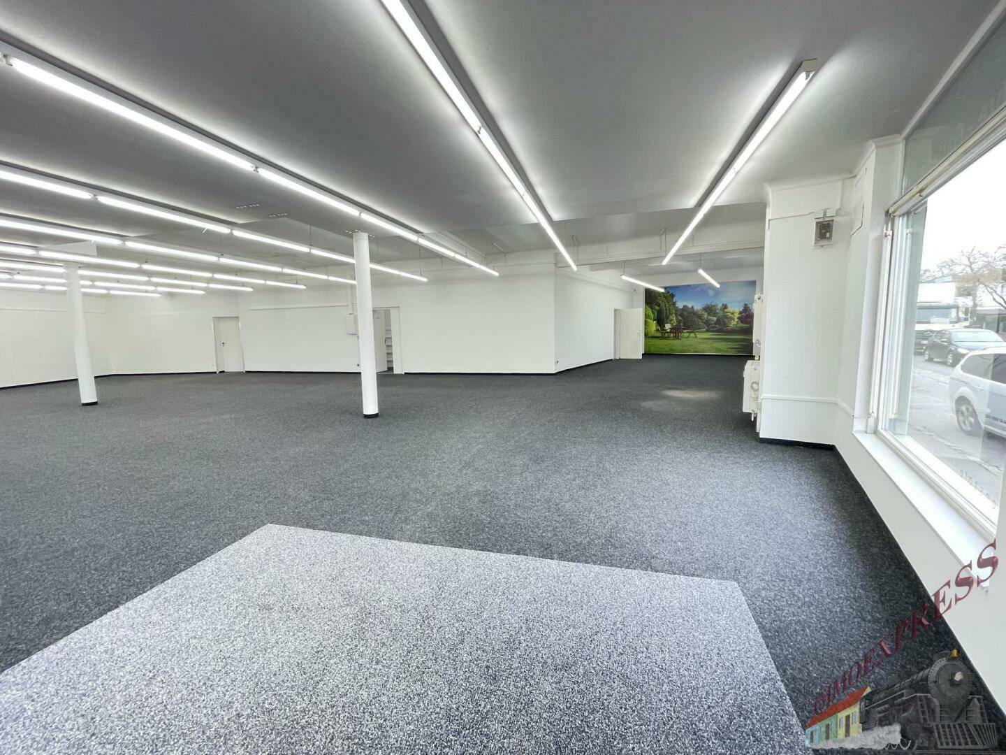 Geräumiger Verkaufsraum mit Teppichboden, großen Fenstern und einer Wandgestaltung mit Landschaftsmotiv.