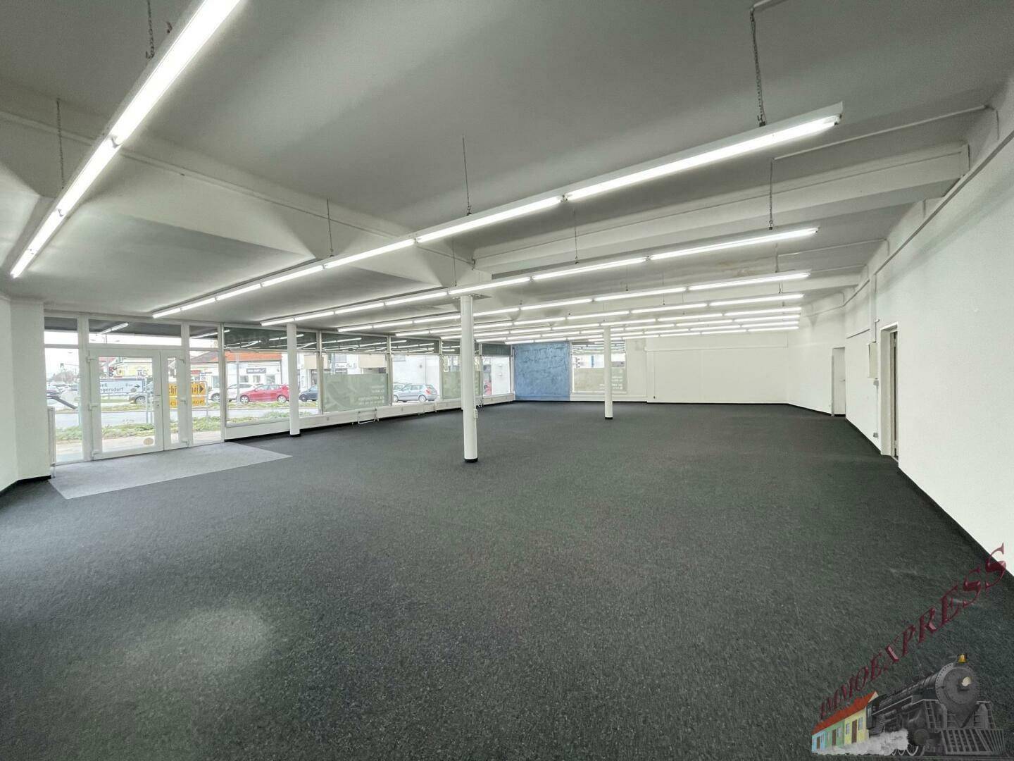 Heller, geräumiger Verkaufsraum mit Teppichboden, Säulen und großen Fensterfronten zur Straße.