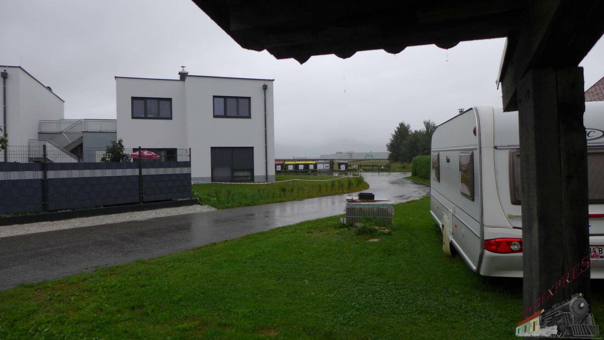 Modernes weißes Haus mit gepflasterter Zufahrt und Wohnwagen auf dem Grundstück an einem regnerischen Tag.