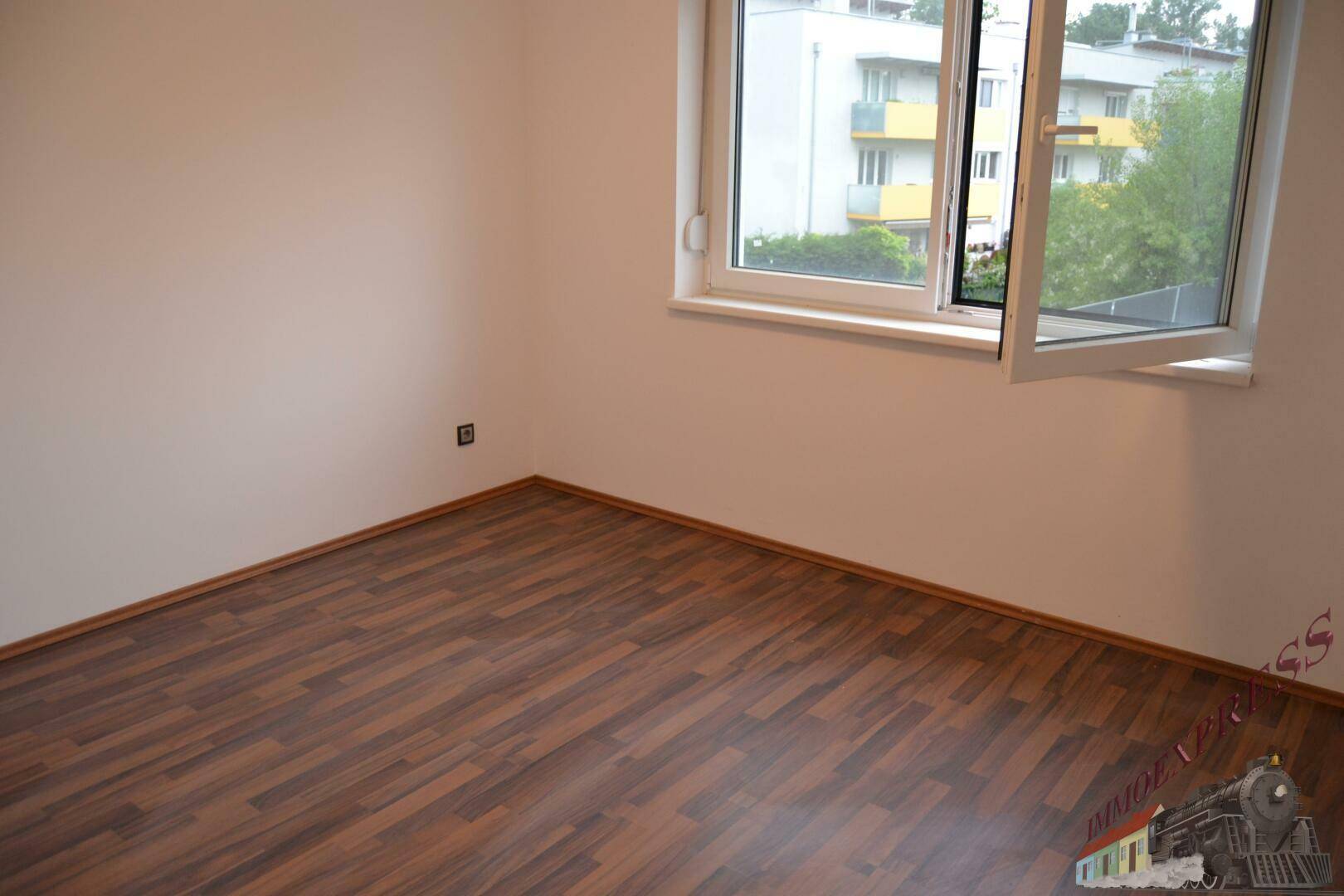 Leeres Zimmer mit dunklem Laminatboden und einem großen Fenster, das den Raum erhellt.