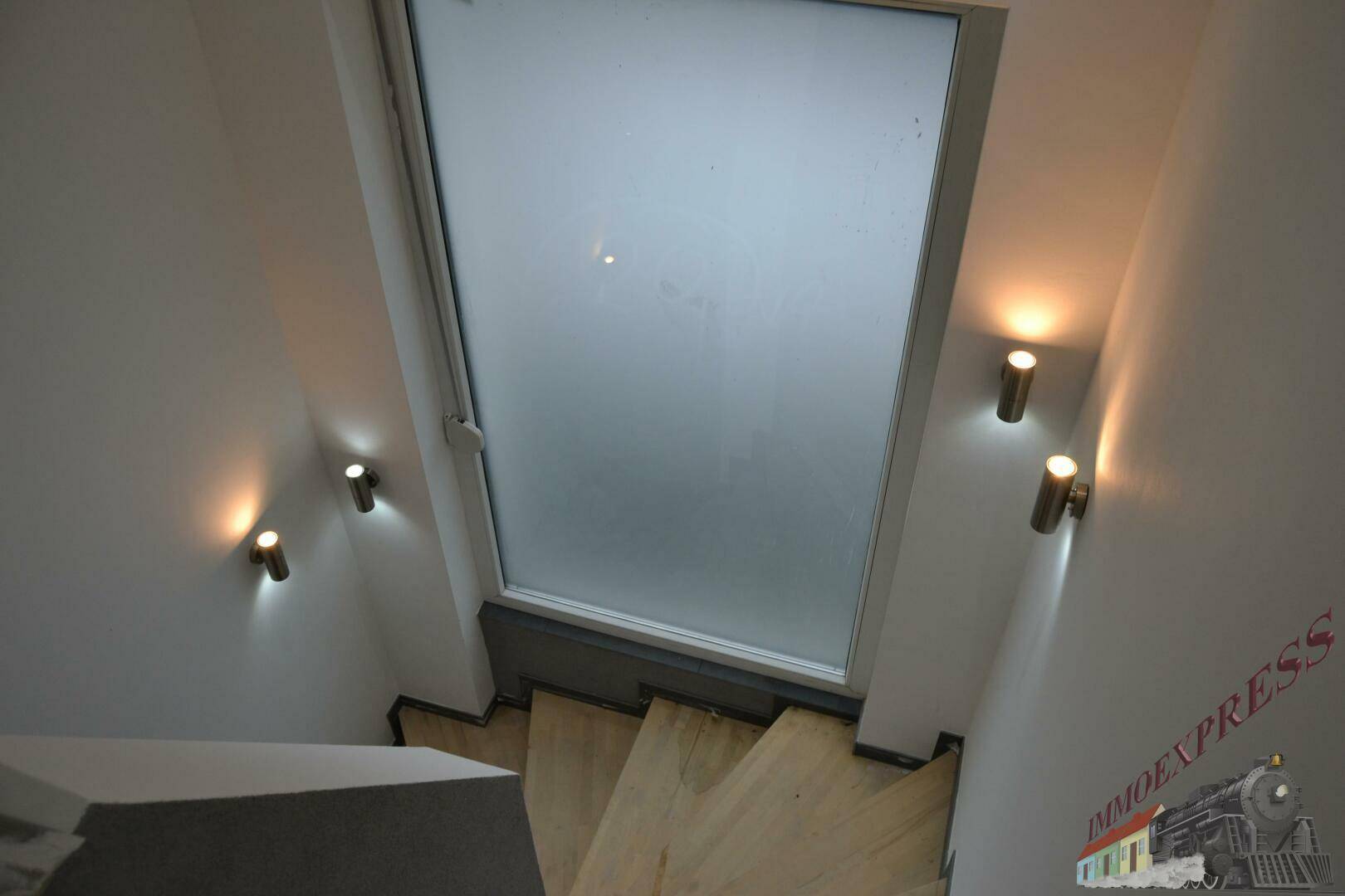 Helle Holztreppe mit Wandleuchten und einem großen Oberlicht, das Tageslicht spendet.