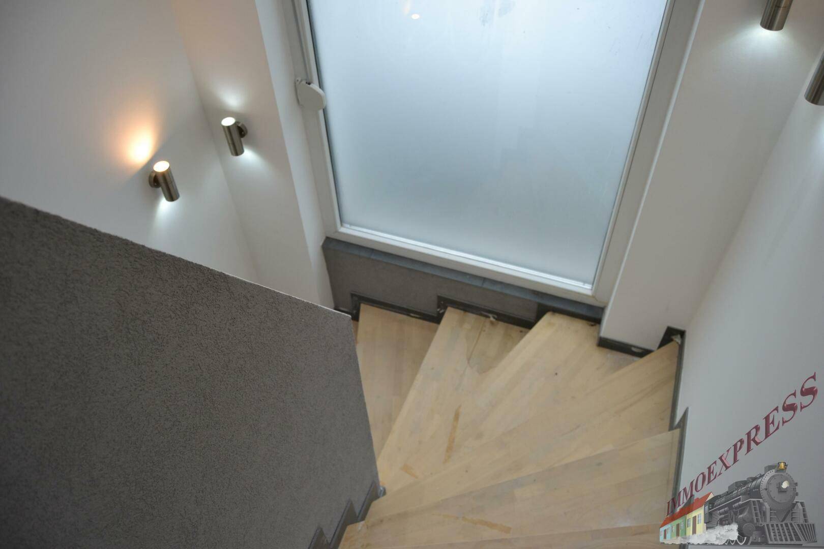 Holztreppe mit Oberlicht und modernen Wandleuchten, die den Aufgang erhellen.