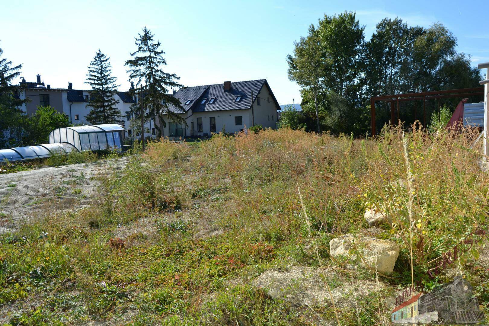 Unbebautes Grundstück mit hohem Gras und Bäumen, im Hintergrund ein Wohnhaus mit Poolüberdachung.
