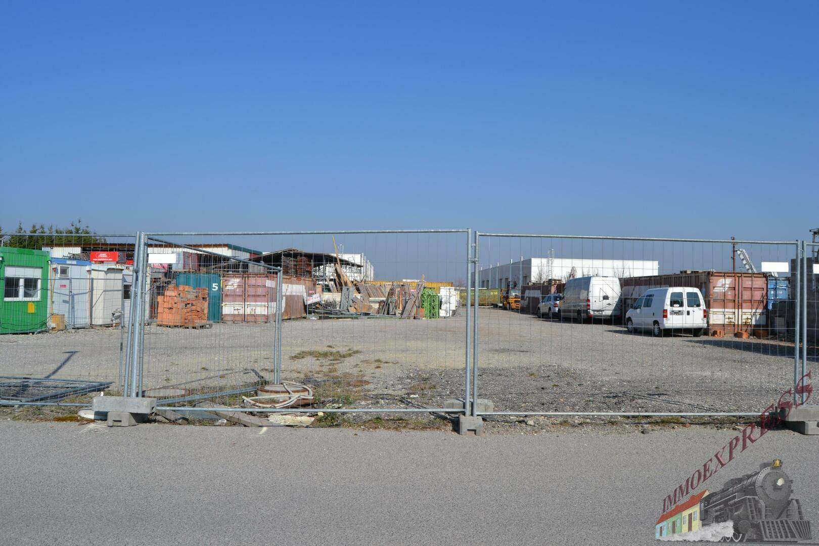 Gewerbefläche mit Bauzaun im Vordergrund, Lagercontainern und Fahrzeugen im Hintergrund.