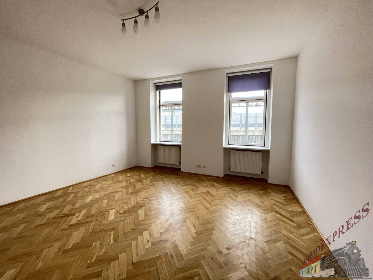 Geräumiges Wohnzimmer mit hellem Fischgrätparkett und zwei großen Fenstern.