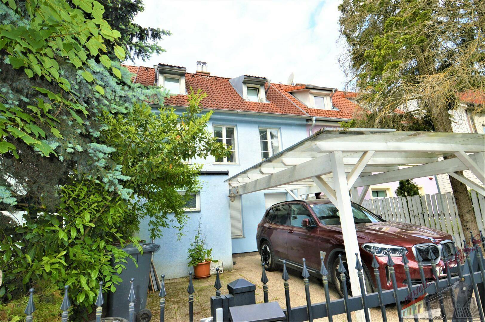Vorderansicht des Hauses mit Carport und Einfahrt, umgeben von Bäumen und Sträuchern.