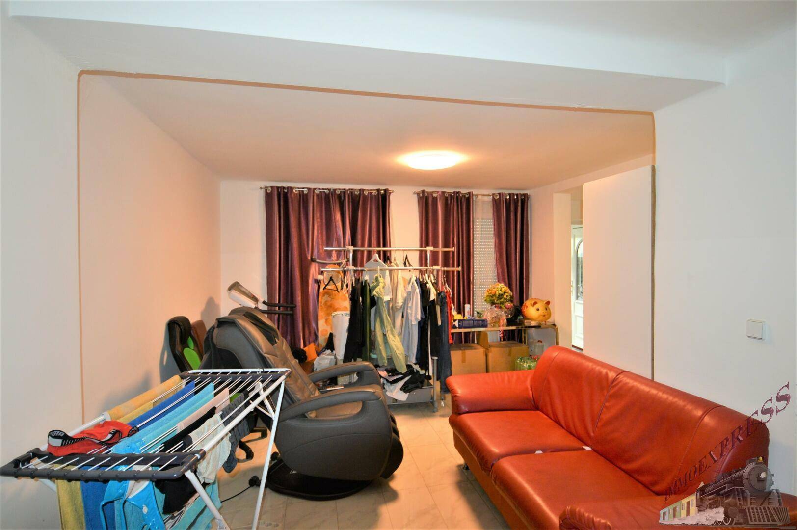 Geräumiges Wohnzimmer mit roten Sofas, Fliesenboden und Blick auf die Fenster mit Vorhängen.