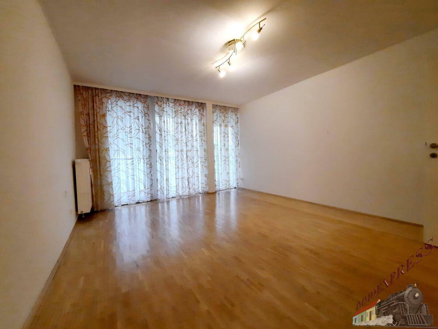 Geräumiges Wohnzimmer mit Parkettboden und Vorhängen an den großen Fenstern.