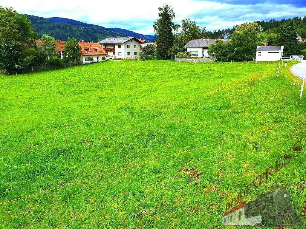 Weitläufige grüne Wiese mit Blick auf mehrere Wohnhäuser und die umliegende Berglandschaft.
