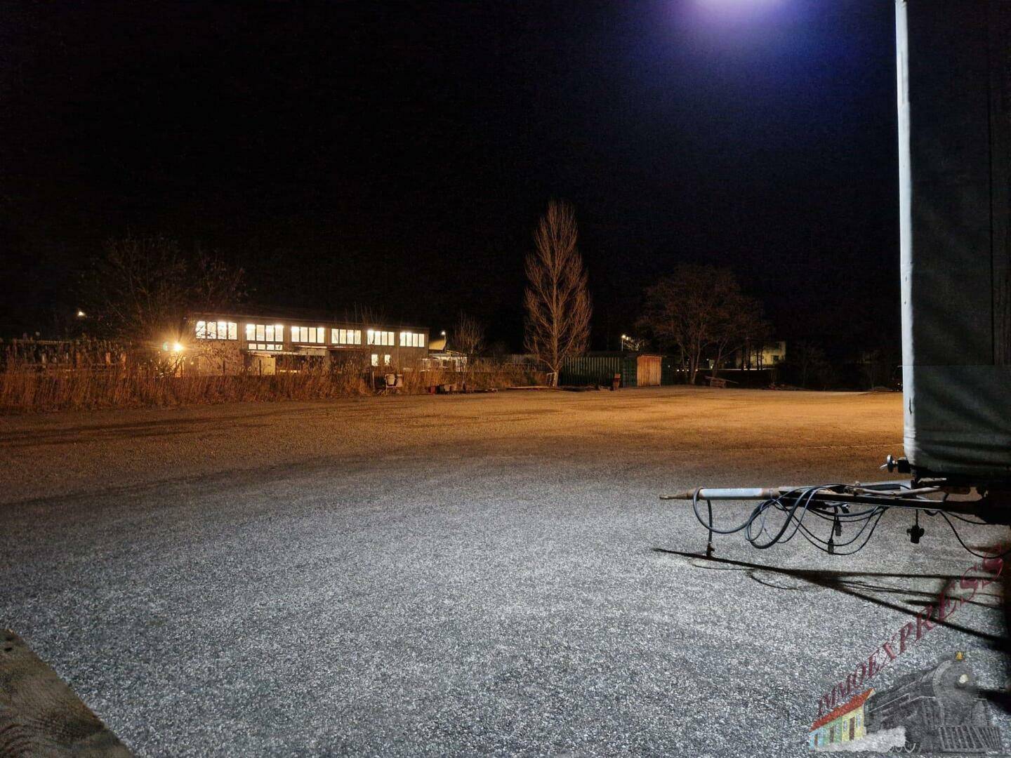 Weitläufiger Schotterplatz bei Nacht mit beleuchtetem Gebäude und hohem Lichtmast im Vordergrund.