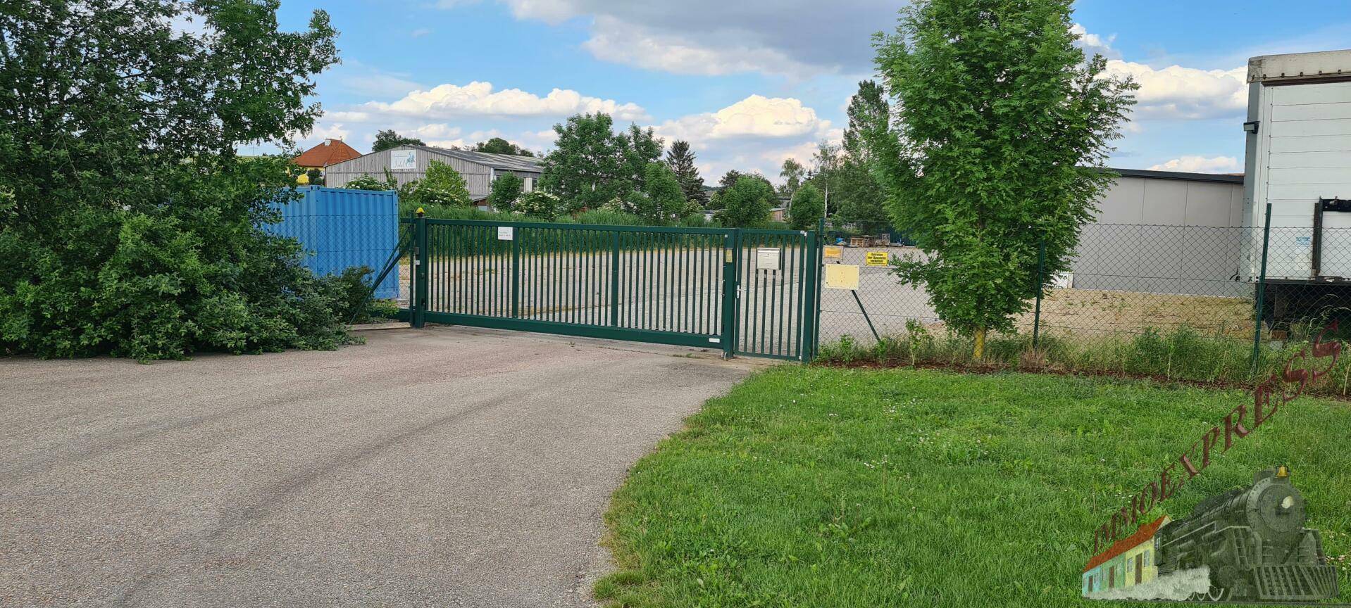 Grünes Metalltor an einer Zufahrt mit Rasenflächen und Industriegebäuden im Hintergrund.