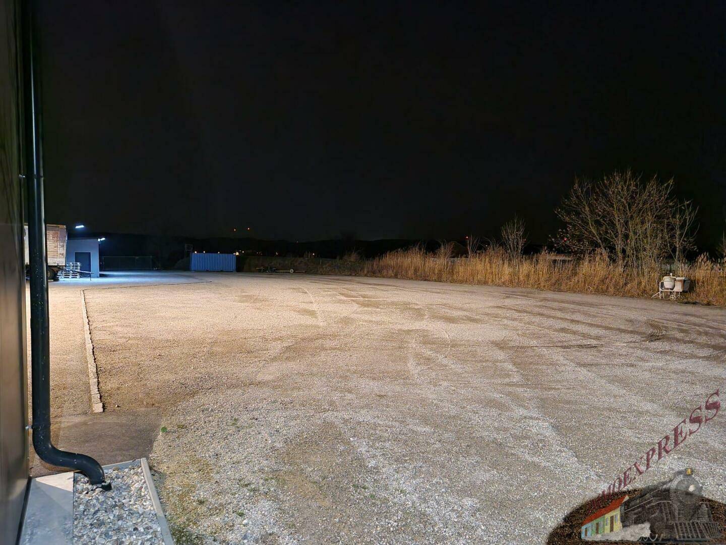 Ausgedehnter Schotterplatz bei Nacht, angrenzend an ein Gebäude mit Regenrinne, unter dunklem Himmel.