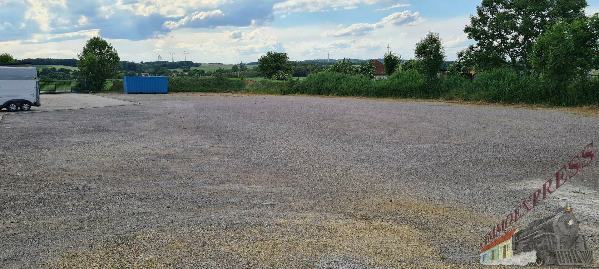 Großer Schotterparkplatz unter bewölktem Himmel, umgeben von Grünflächen und Hügeln in der Ferne.