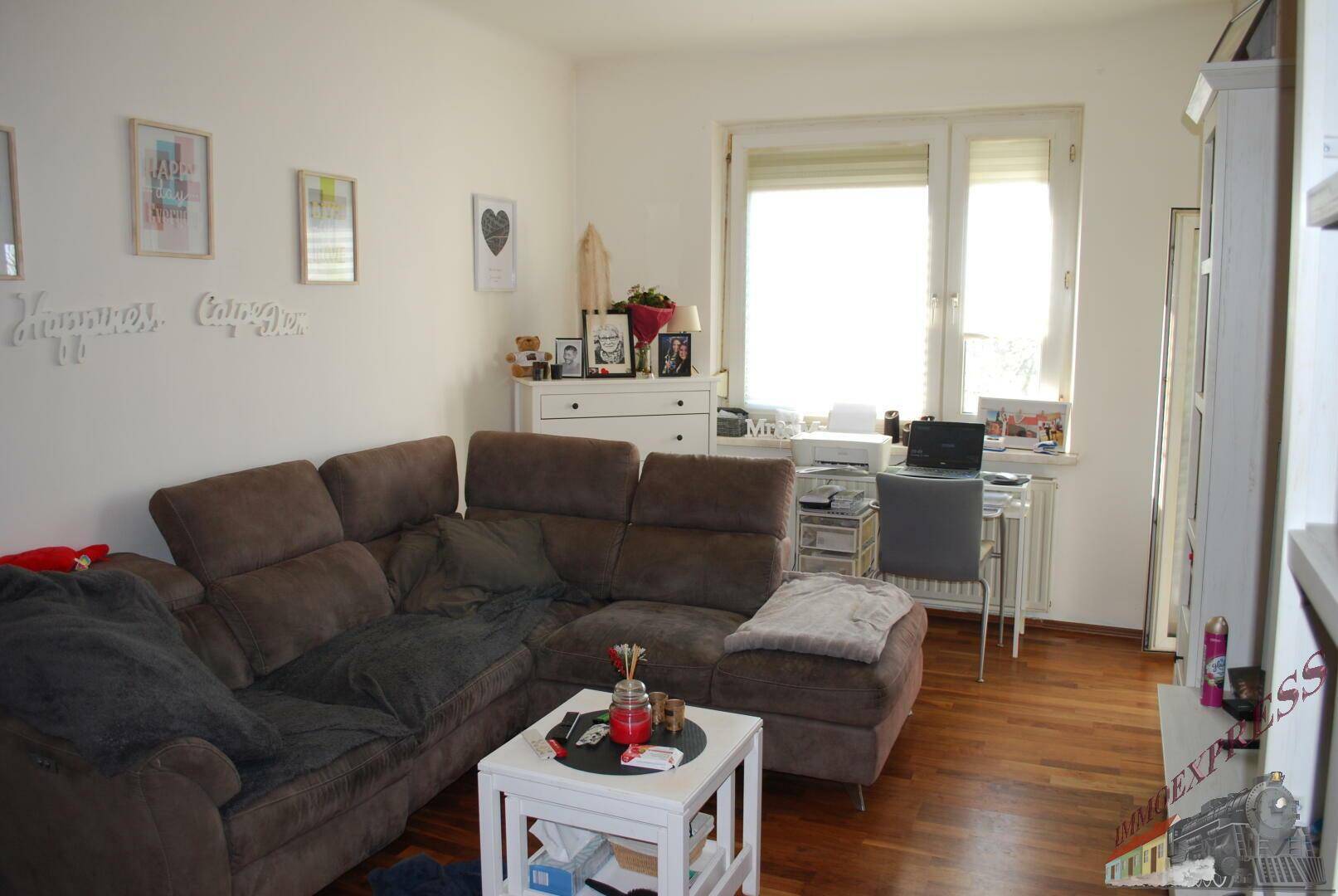 Gemütliches Wohnzimmer mit großem Ecksofa, Holzboden und einem hellen Arbeitsbereich am Fenster.
