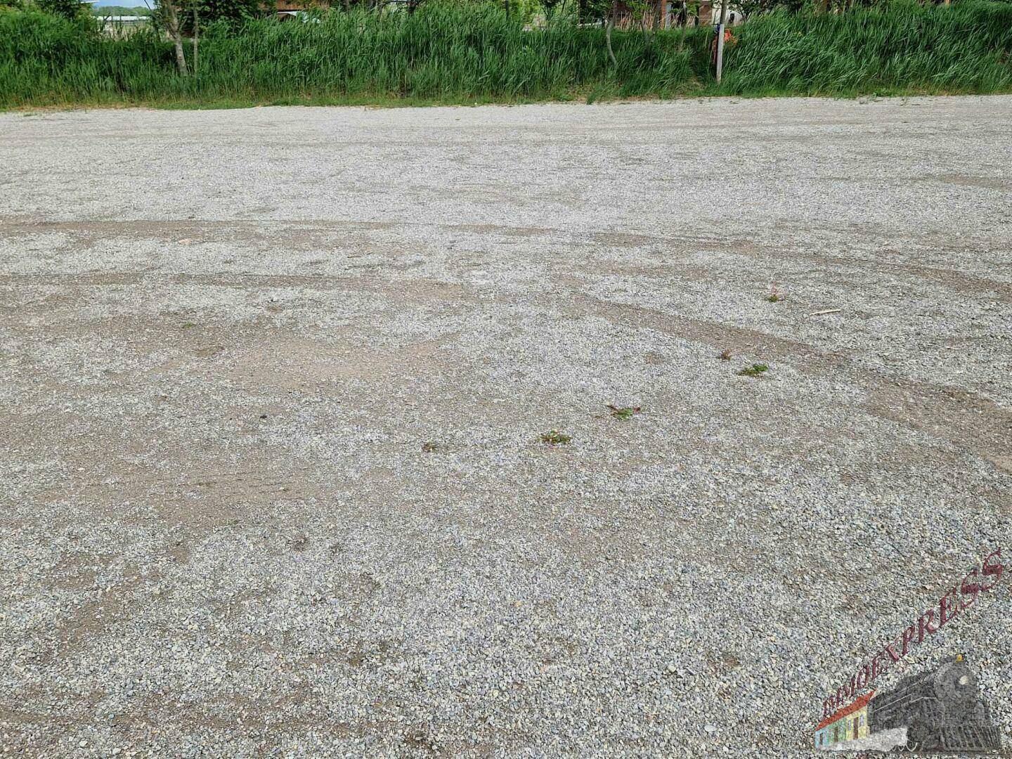 Weitläufiger, leerer Schotterplatz mit vereinzelten Grashalmen und einer grünen Hecke im Hintergrund.