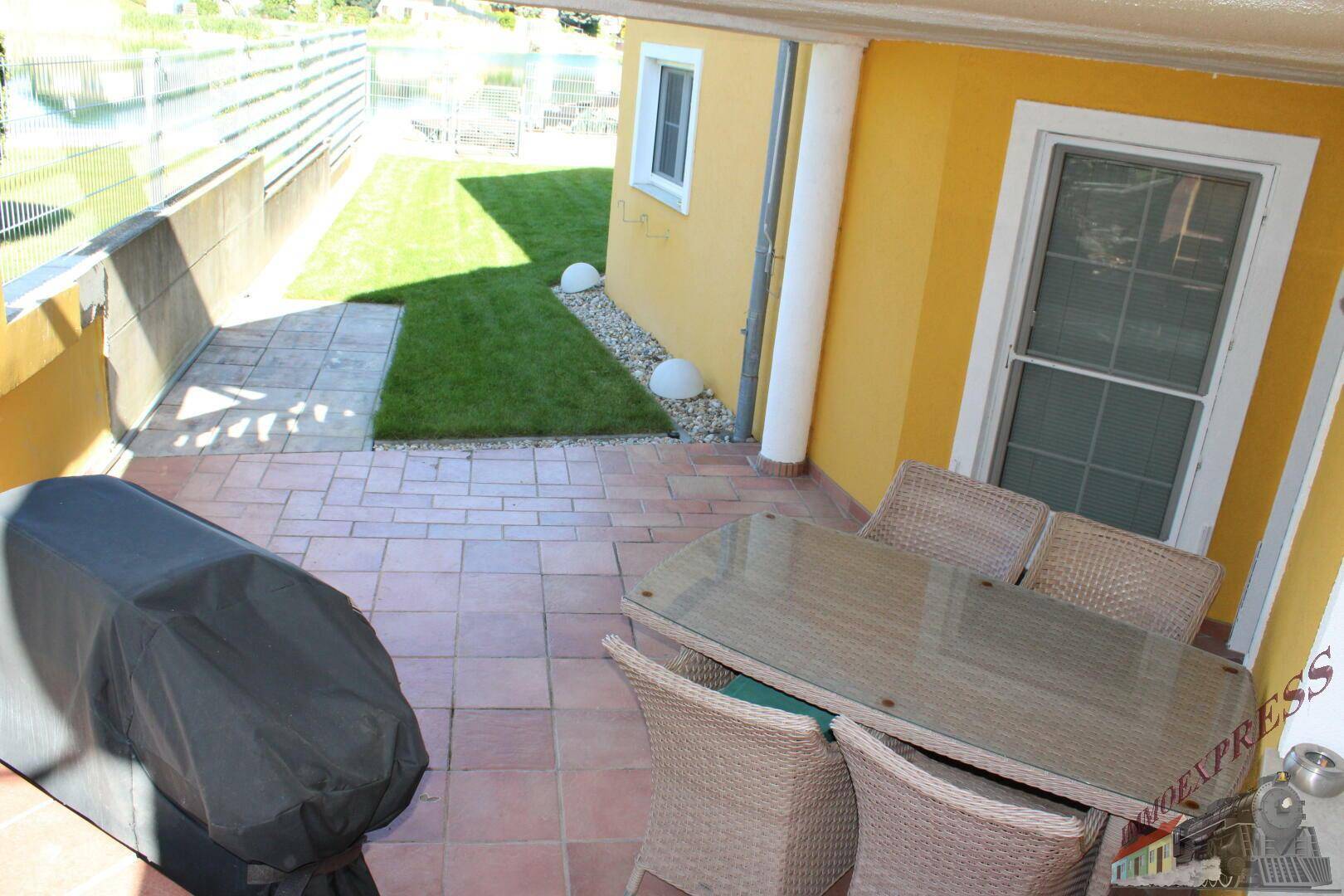 Überdachte Terrasse mit Fliesenboden und Gartenmöbeln, angrenzend an eine kleine Rasenfläche.