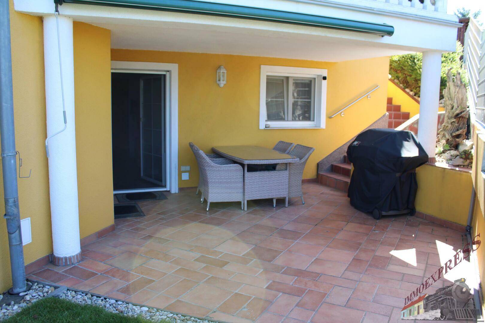 Überdachte Terrasse mit Fliesenboden, Esstisch und Grillbereich am gelben Haus.