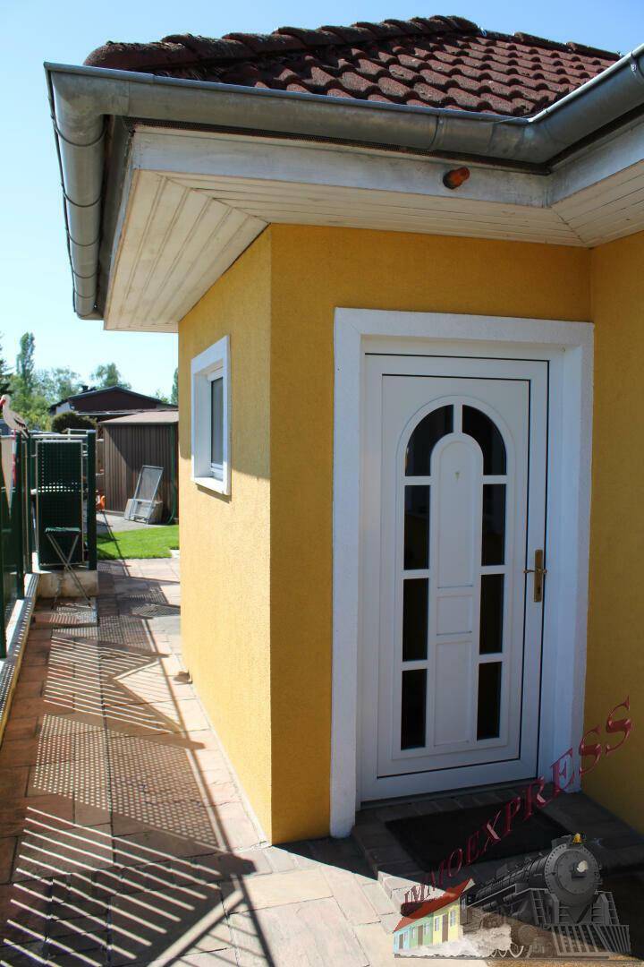 Gelbe Fassade eines Nebengebäudes mit weißer Eingangstür und braunem Ziegeldach.