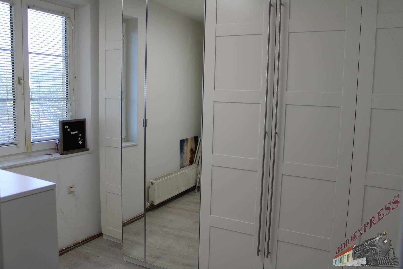 Schlafzimmer mit weißem Kleiderschrank, Spiegeltür und Fenster mit Jalousien.