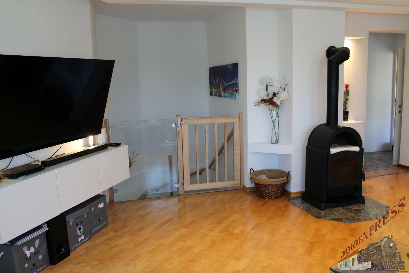 Wohnzimmer mit Holzboden, Fernseher und einem gemütlichen Holzofen, Treppenzugang.