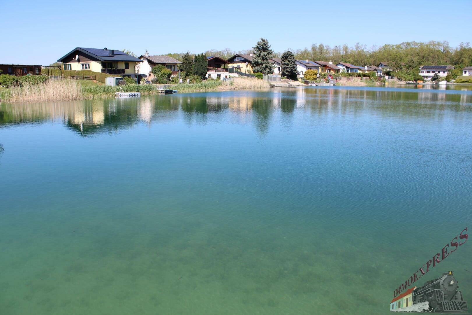 Weitläufiger Blick über einen klaren See mit Häusern am gegenüberliegenden Ufer unter blauem Himmel.