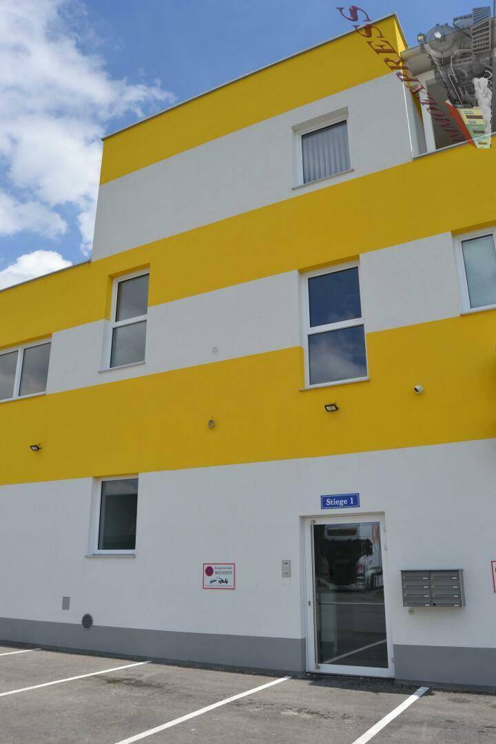 Eingangsbereich eines modernen Gebäudes mit gelb-weißer Fassade und Parkplätzen davor.