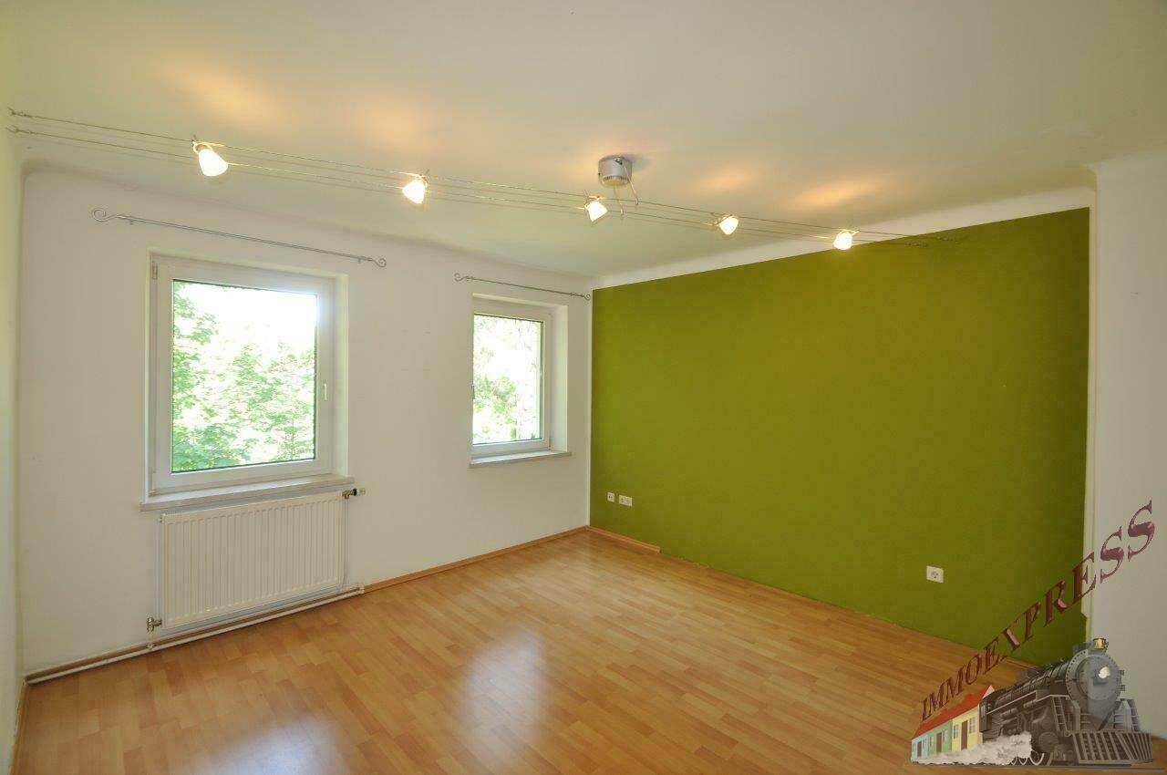 Helles Wohnzimmer mit grüner Akzentwand, Holzparkettboden und zwei Fenstern.