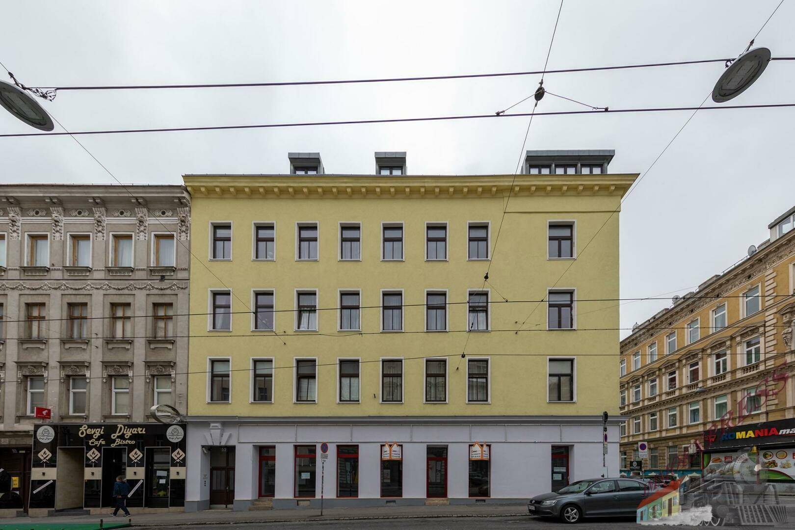 Gelbes Mehrfamilienhaus an einer belebten Straße mit vielen Fenstern und Geschäften im Erdgeschoss.