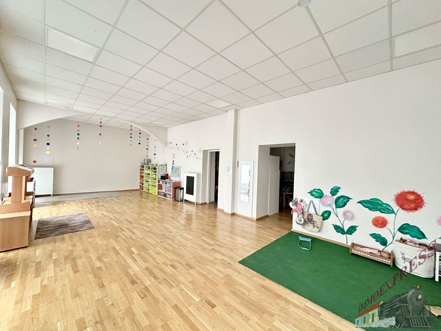 Geräumiger und heller Spielraum mit Holzboden, farbenfrohen Wandmalereien und einer grünen Teppichfläche.