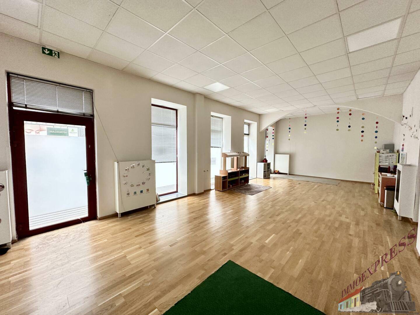 Heller und weitläufiger Spielbereich mit Holzboden, großen Fenstern und einer grünen Teppichzone, gut beleuchtet.