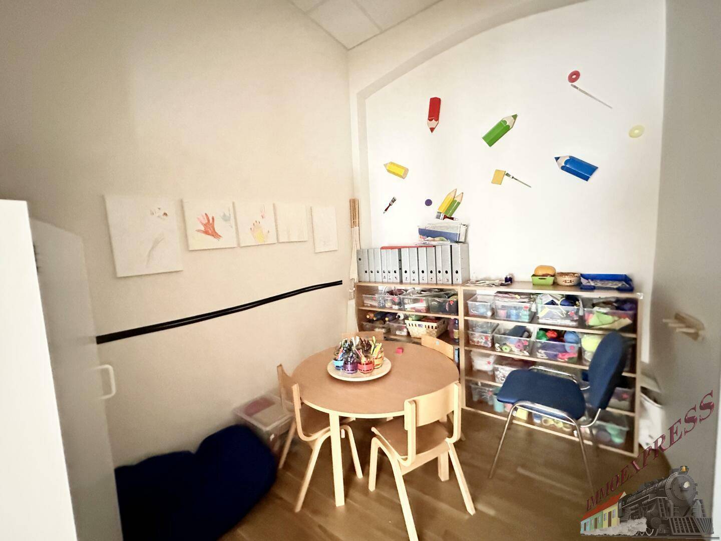 Kleiner, gemütlicher Aktivitätsraum mit Holztisch und Stühlen, Regalen und farbenfroher Wanddekoration für Kinder.