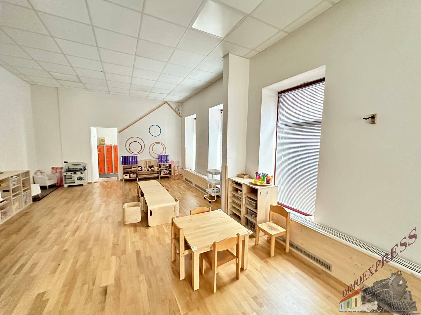 Heller und geräumiger Spielraum mit Holzboden, kleinen Holztischen und Stühlen, gut beleuchtet durch Fenster.