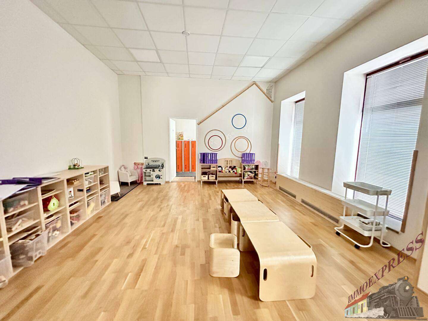 Geräumiger Spielraum mit Holzboden, kleinen Holztischen und einer Wickelstation, gut beleuchtet und funktional.