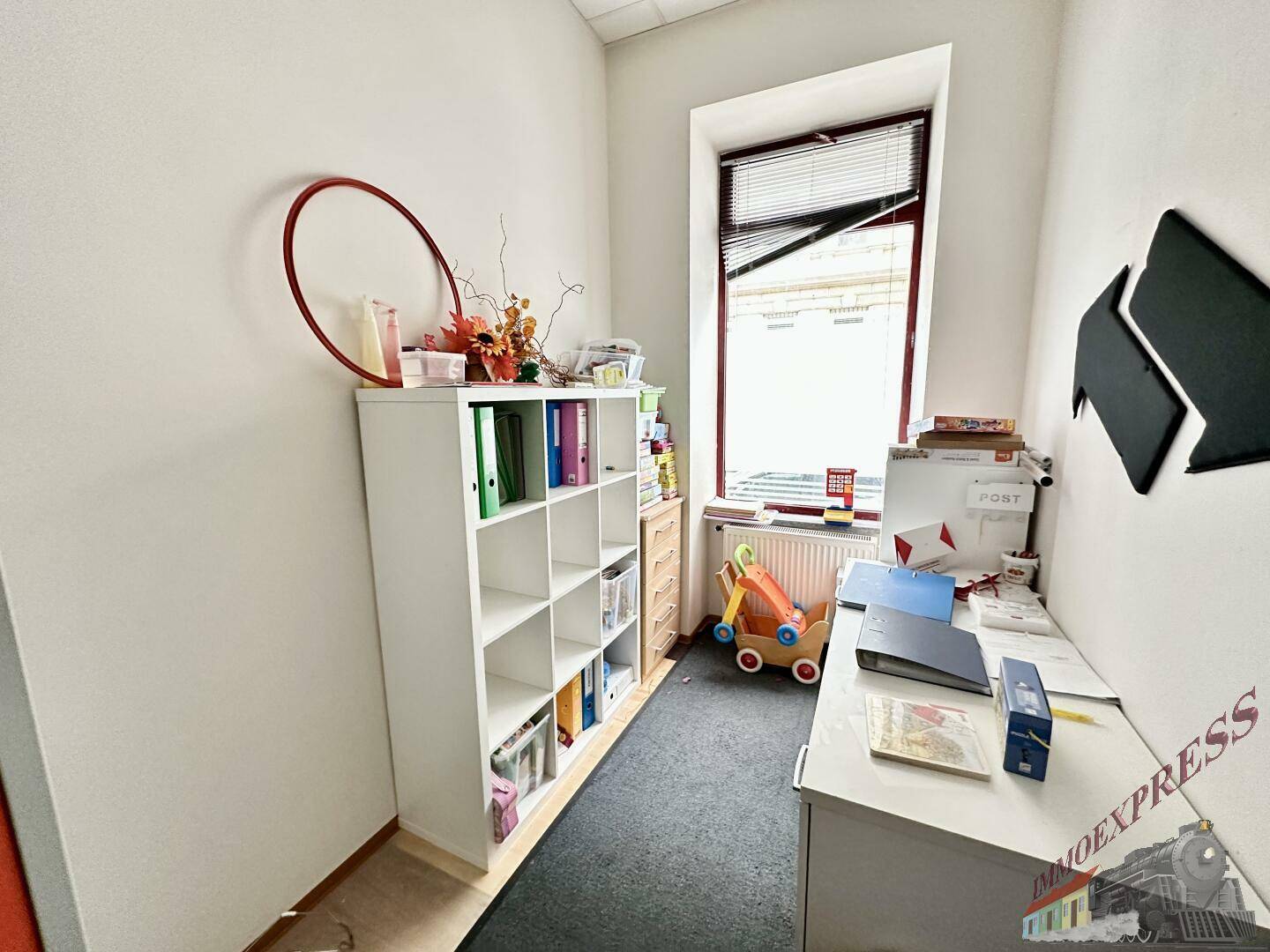 Kleiner Bürobereich mit Schreibtisch, Regal und Fenster, ideal für administrative Aufgaben oder als Arbeitszimmer.