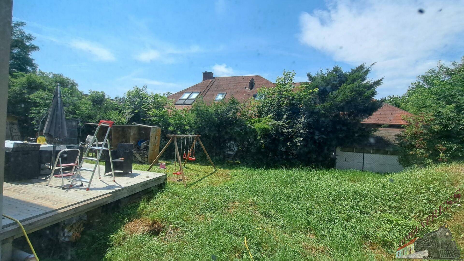Grüner Garten mit Holzterrasse, Schaukel und Blick auf Nachbargebäude.