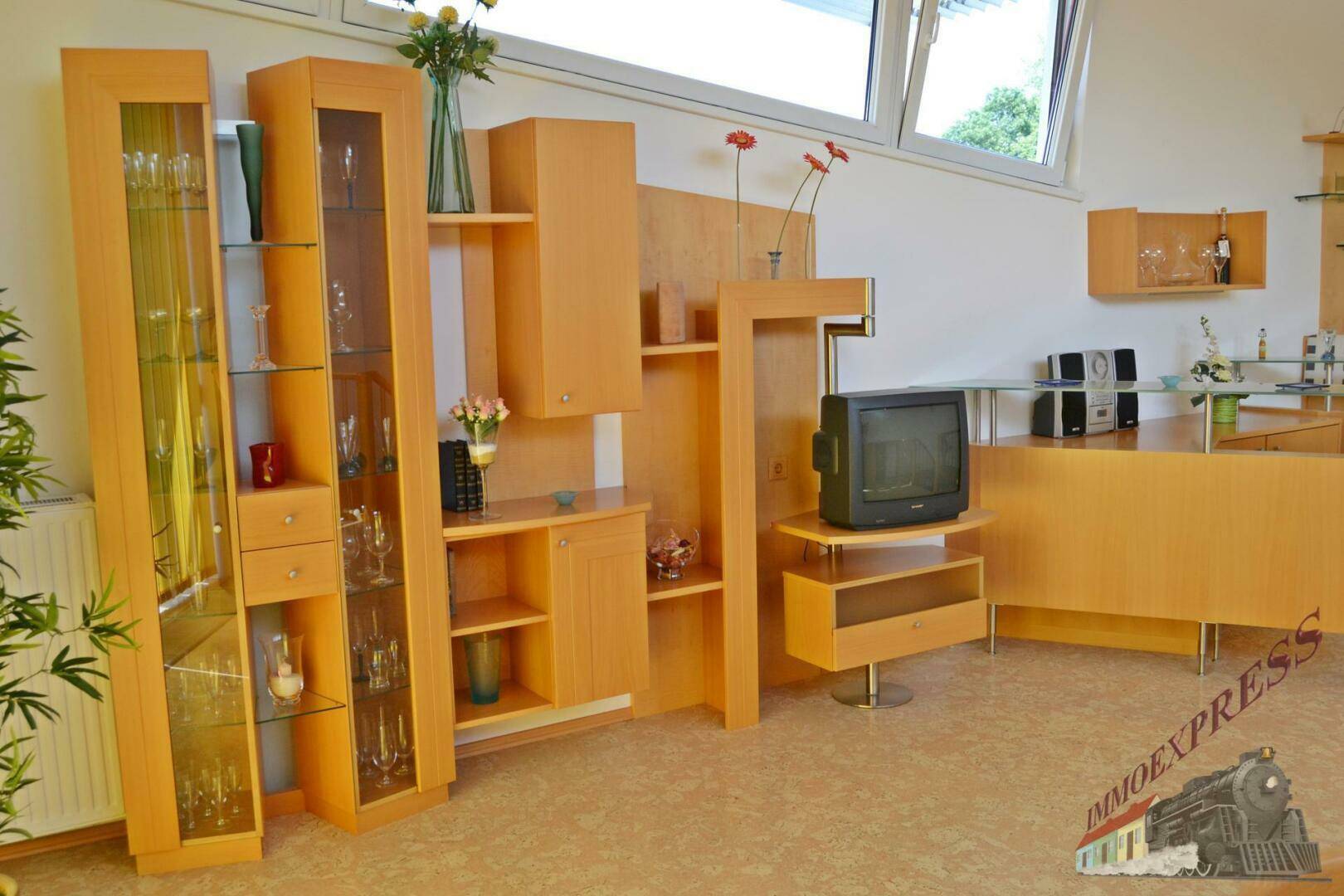 Wohnbereich mit hellen Holzmöbeln, Vitrinen und einem älteren Fernseher, gut gepflegt und funktional.