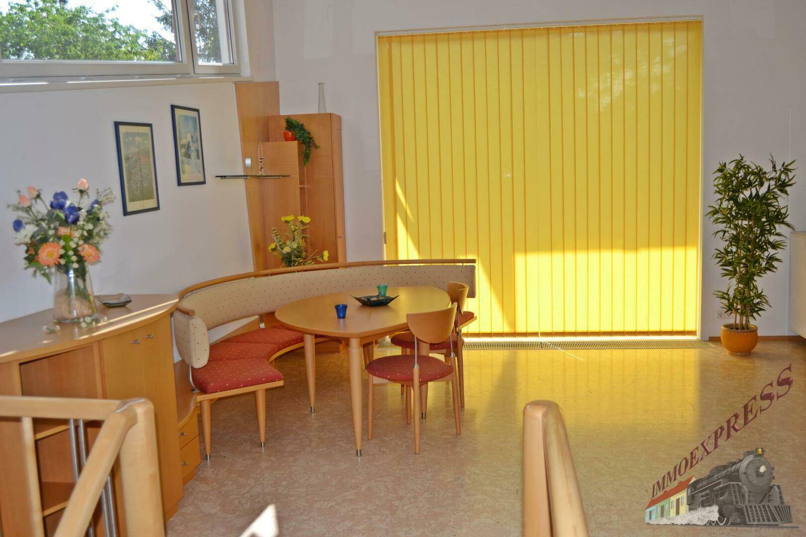 Heller Essbereich mit runder Eckbank, Holztisch und großen Fenstern mit gelben Vertikaljalousien.