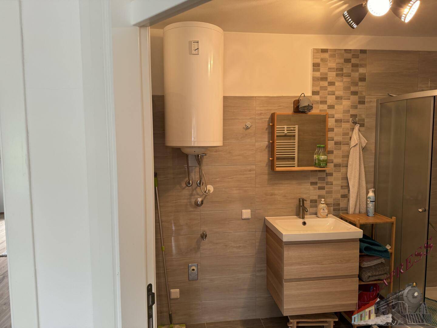 Badezimmer mit Waschtisch, Spiegel und Warmwasserboiler, funktional und hell gefliest.