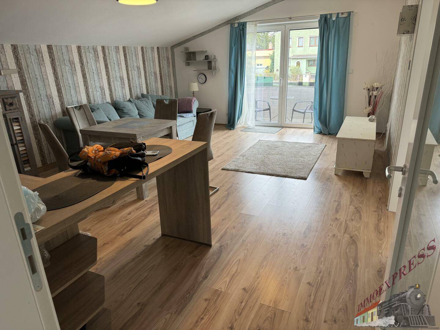 Heller Wohn- und Essbereich mit Holzboden, Zugang zum Balkon und blauen Vorhängen.