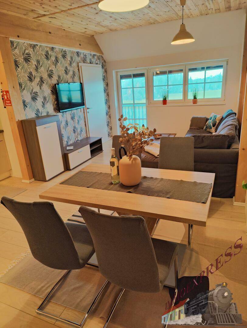 Gemütlicher Wohn-Essbereich mit Holzbalken, Esstisch und Sofa, mit Blick auf die grüne Landschaft durch die Fenster.