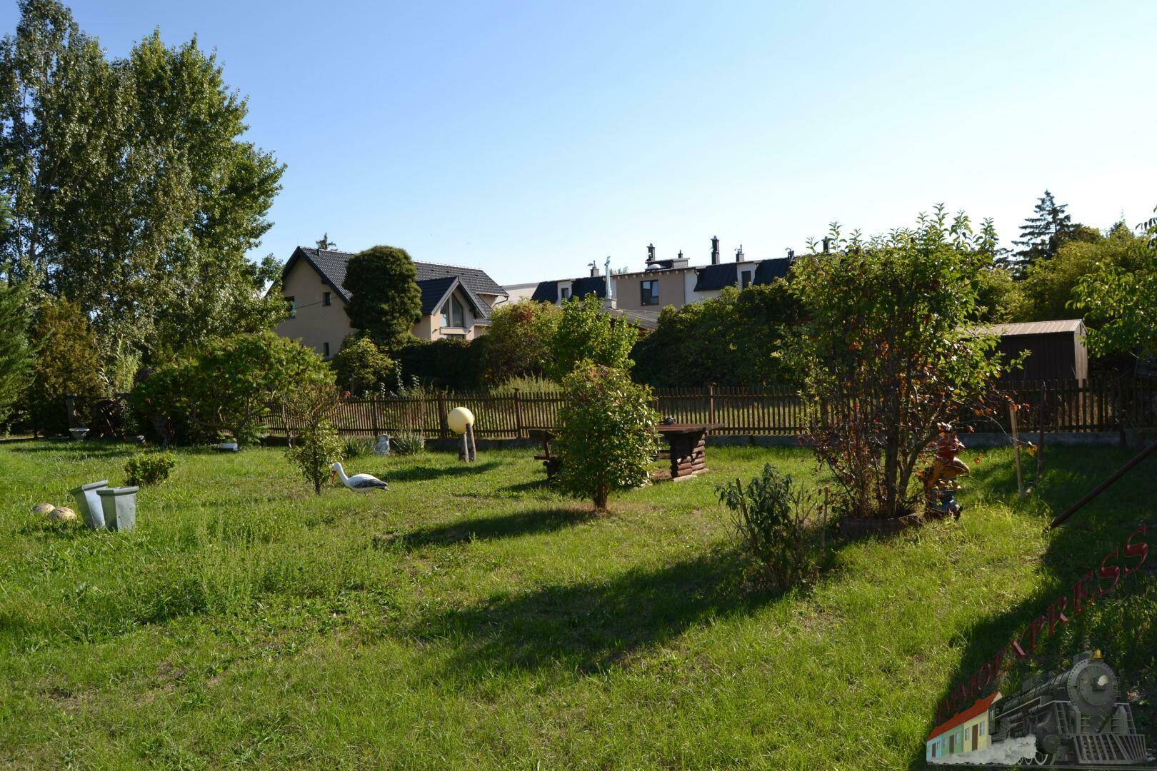 Gepflegter Gartenbereich mit Rasenfläche, Bäumen und einem Grillplatz im Freien unter blauem Himmel.