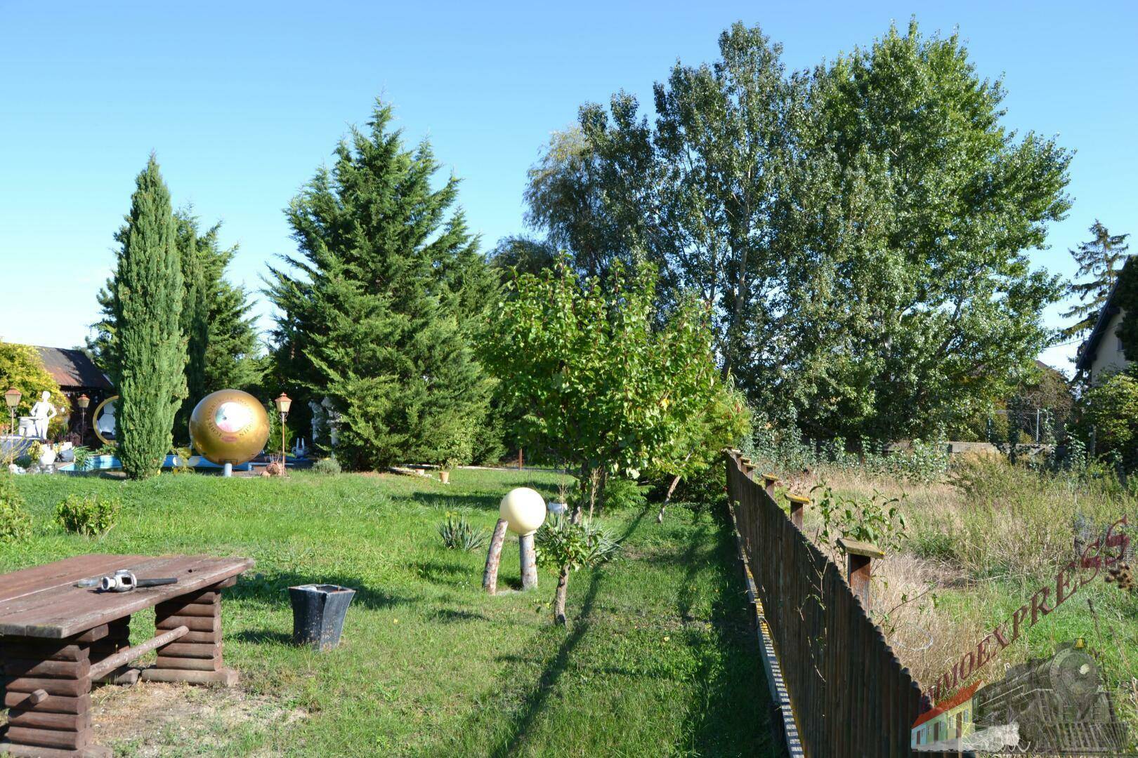 Sonniger Garten mit Holztisch, verschiedenen Bäumen und dekorativer goldener Kugel unter blauem Himmel.