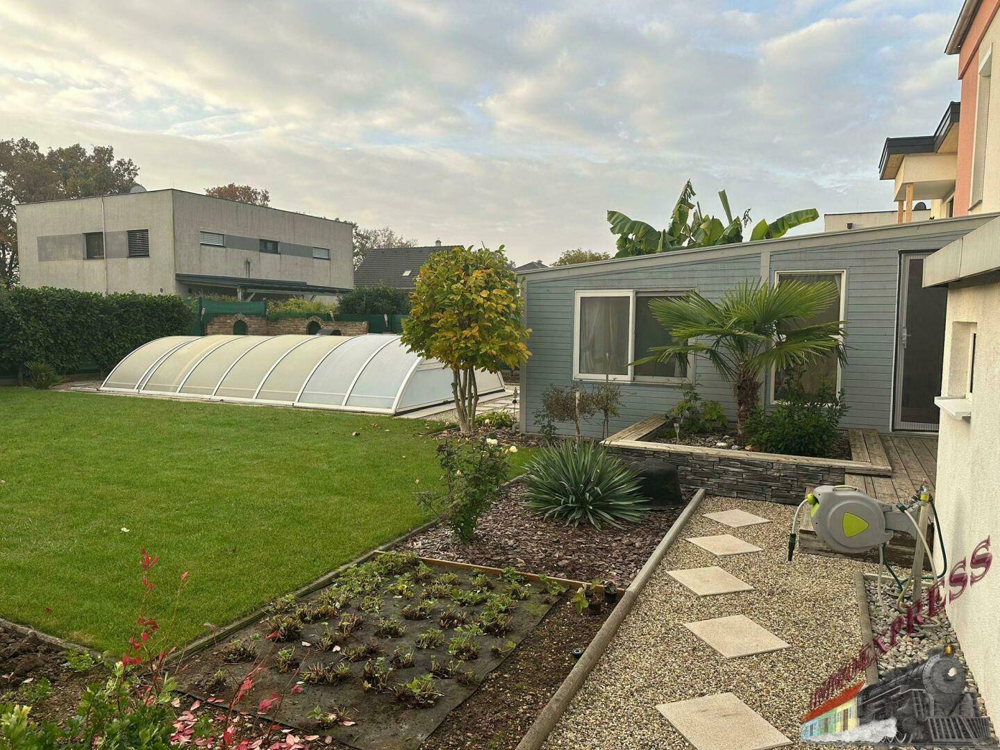 Weitläufiger Garten mit Rasen, bepflanzten Beeten, Wintergarten und überdachtem Poolbereich.