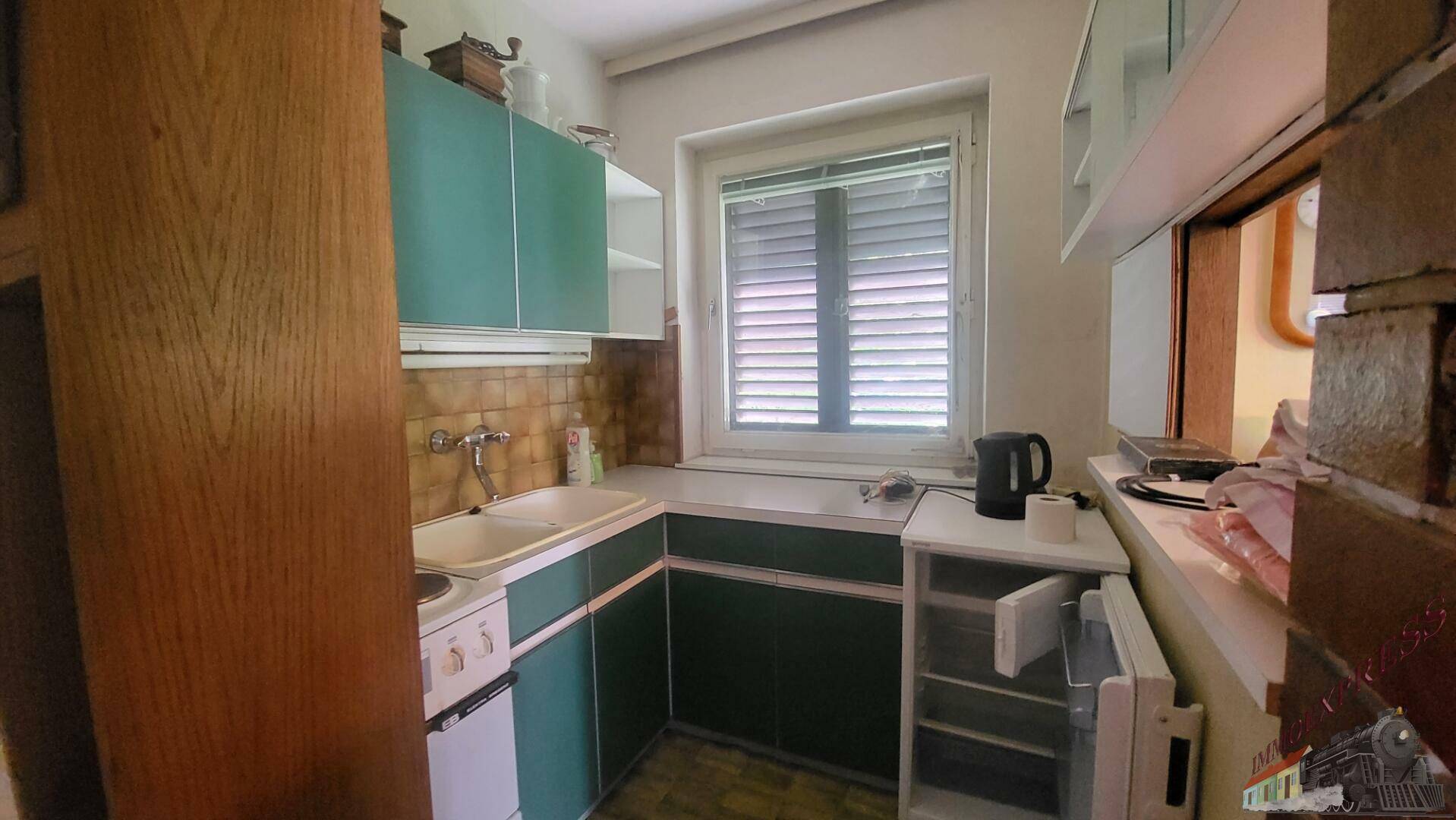 Kleine Küche mit tealfarbenen Schränken, Spüle und Fenster mit Jalousien.