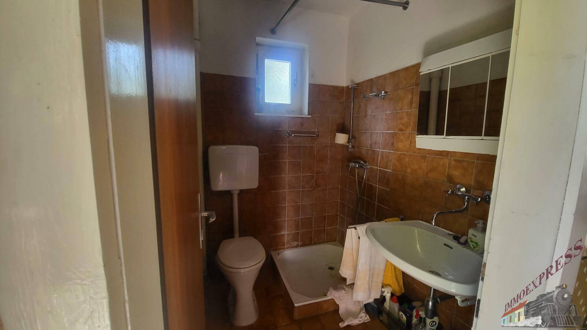 Badezimmer mit braunen Fliesen, Dusche, WC und kleinem Fenster für Belüftung.