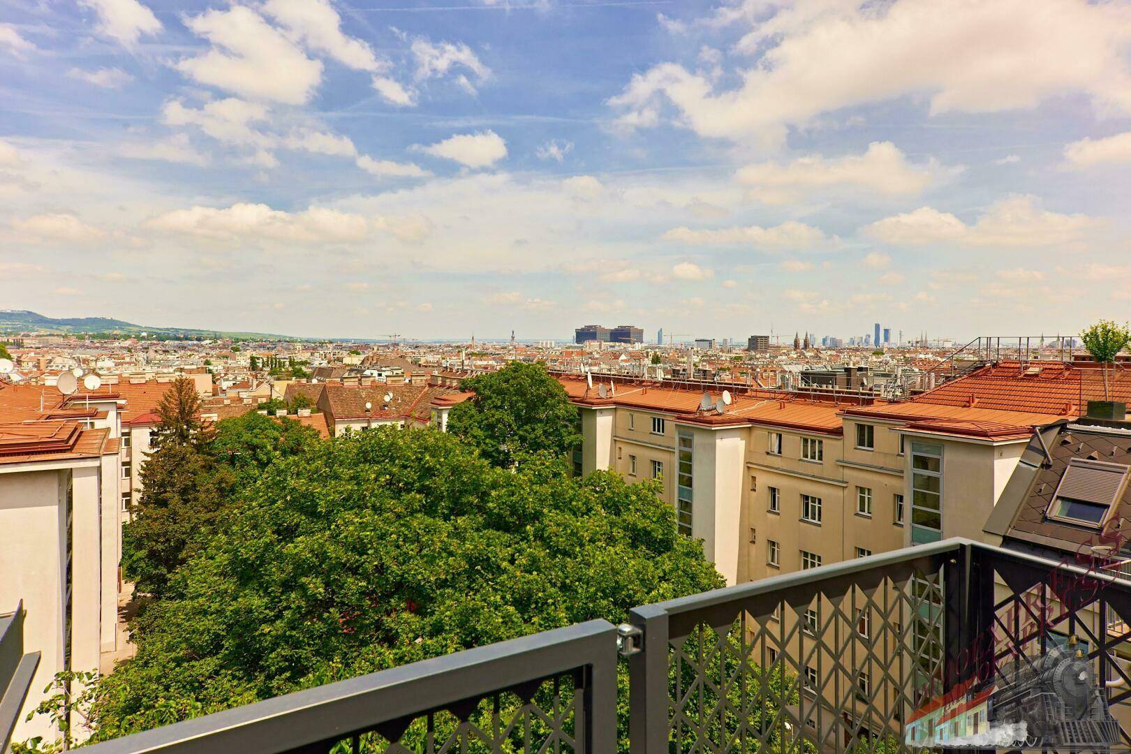 Weitläufiger Blick über die Stadt mit grünen Bäumen und roten Dächern unter blauem Himmel.