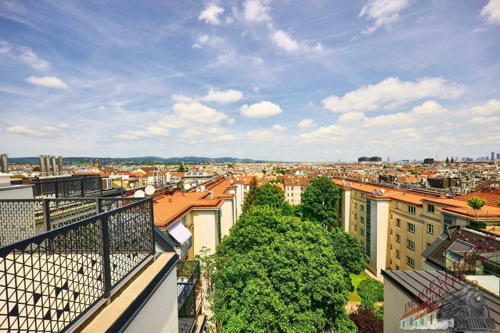 Weitläufiger Blick über die Stadt mit grünen Bäumen und roten Dächern unter blauem Himmel.